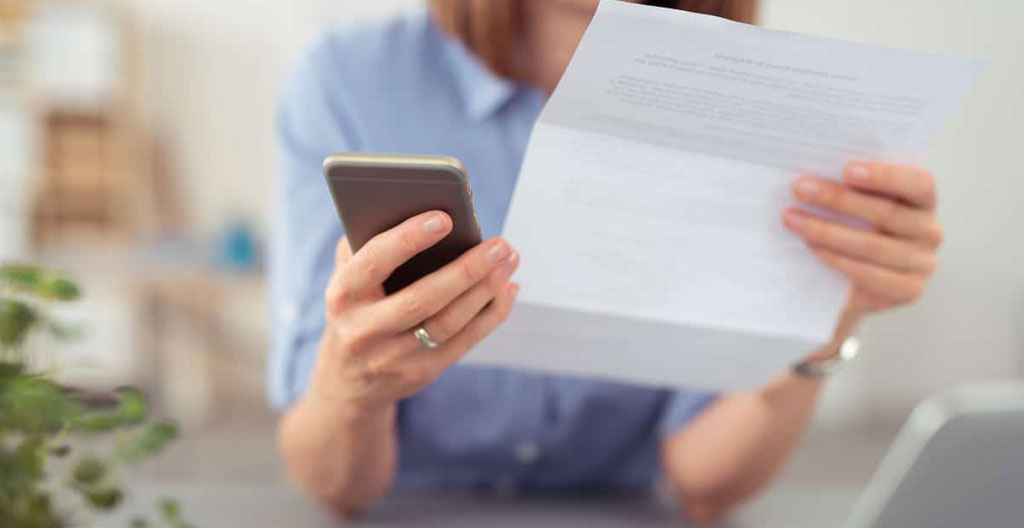 Mains tenant une carte d'identité française et un justificatif de domicile EDF sur un bureau blanc avec ordinateur portable en arrière-plan